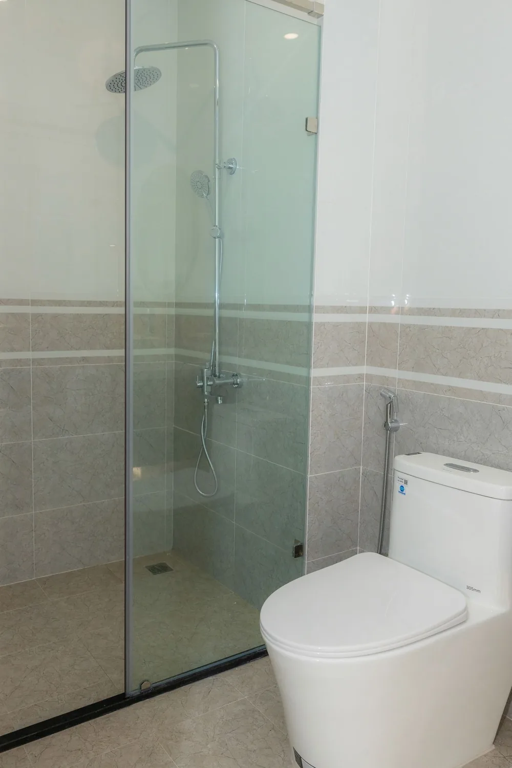 Pancuran kaca dan kloset dalam kamar mandi terang